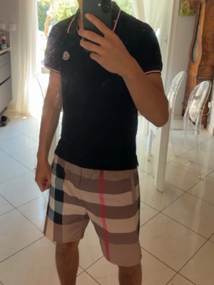 Svart Moncler t-shirt med röd/vit kant - Svart t-shirt från Moncler med korta ärmar och klassisk polokrage. Röd och vit randig kant på krage och ärmslut ger en clean och sportig vibe. Moncler-logga broderad på bröstet. Tillverkad i mjuk bomull för skön komfort.