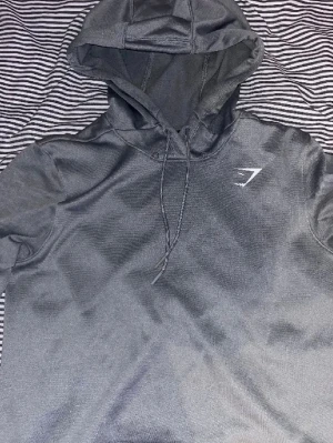 Mörkgrå hoodie från Gymshark - Snygg mörkgrå hoodie från Gymshark med dragsko i huvan och diskret logga på bröstet. Tillverkad i ett mjukt och stretchigt material som passar perfekt till både träning och chill. Klassisk passform med långa ärmar och ribbade muddar. 