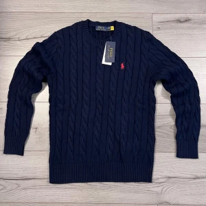 Polo Ralph Lauren kabelstickad tröja mörkblå  - Kabelstickad tröja i mörkblått från Polo Ralph Lauren med rund hals och röd broderad logga på bröstet. Tröjan har ribbade muddar och är tillverkad i mjukt material som ger en klassisk och stilren look.