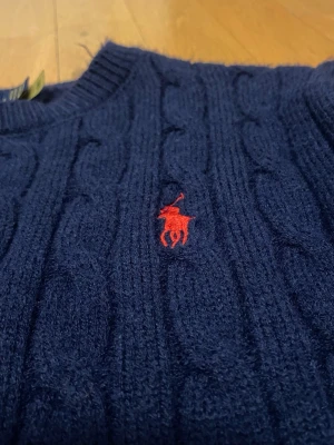 Mörkblå stickad tröja från Polo Ralph Lauren - Mörkblå kabelstickad tröja från Polo Ralph Lauren med rund halsringning och röd broderad logga på bröstet. Långa ärmar och ribbstickade muddar vid ärmslut och nederkant. Perfekt för lager-på-lager och klassisk stil.