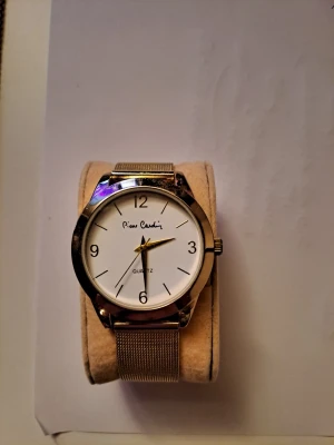Guldfärgad klocka från Pierre Cardin - Stilren klocka från Pierre Cardin med vit urtavla och guldfärgat mesh-armband i metall. Klassisk rund form med tydliga svarta siffror och tunna visare. Perfekt accessoar för dig som gillar enkel och elegant design.