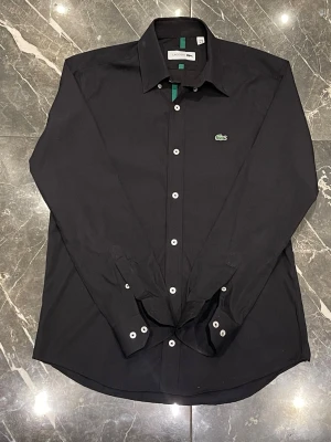 Svart skjorta från Lacoste - Klassisk svart skjorta från Lacoste med vita knappar och den ikoniska gröna krokodilloggan på bröstet. Skjortan har lång ärm, normal passform och är tillverkad i mjuk bomull. Perfekt för en clean och stilren look. Aldrig använd!