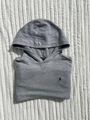 Grå hoodie från Polo Ralph Lauren - Snygg grå hoodie från Polo Ralph Lauren med klassisk huva och liten broderad logga på bröstet. Hoodien har känguruficka framtill och ribbade muddar vid ärmslut och nederkant. Perfekt för en chill och stilren look.