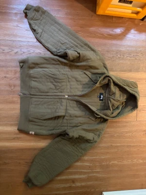 Olivgrön quiltad hoodie från Stussy - Säljer en olivgrön quiltad hoodie från Stussy med dragkedja framtill och två fickor. Hoodien har ribbade muddar och en stor huva. Materialet är tjockt och vadderat, perfekt för kyligare dagar. Stussy-logga på etiketten vid midjan.