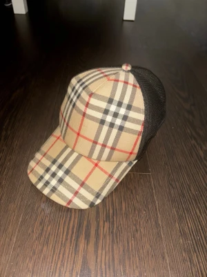 Beige keps med klassiskt rutmönster - Säljer en snygg keps från Burberry med ikoniskt beige, svart och rött rutmönster. Framsidan är i tyg med mönster och baksidan är i svart mesh för extra ventilation. Perfekt för dig som vill ha en trendig accessoar med lyxig känsla.