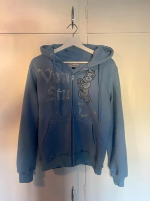Blå vonstudio hoodies - Ljusblå hoodie från Vonstudio med dragkedja, huva och fickor framtill. Dekorerad med strassbokstäver och en söt ängel på bröstet. Perfekt för dig som gillar streetwear och vill sticka ut med detaljer. Tillverkad i mjukt material för extra komfort. Bara att höra av er om ni undrar något. Köpte den ny från vonstudio. Alla diamanter sitter kvar.