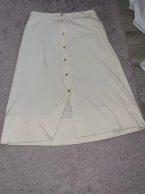 Ljusbeige långkjol med knappar (Storlek 46) - Stilren ljusbeige långkjol från Turkiet med knappar framtill och en liten slits nedtill. Den har praktiska fickor samt en dragkedja placerad bredvid fickorna för en snygg och diskret detalj.  Materialet är ogenomskinligt 🩷