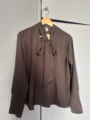 Svart blus med knyt från H&M - Stilren svart blus från H&M med knytband i halsen och långa ärmar. Blusen har en mjuk, något glansig yta och breda manschetter vid ärmsluten. Perfekt för en trendig och elegant look. Prislapp kvar för 299kr.