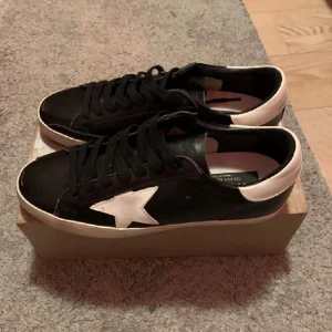 Svarta Golden Goose  - Säljer ett par svarta Golden Goose Supero-Star sneakers med vit stjärndetalj på sidan och vit häl. Skorna har svarta skosnören och vit sula. Klassisk låg modell med cool kontrast mellan svart och vitt. Perfekta för dig som gillar streetstyle och vill sticka ut. Använda 2 gånger vid speciella tillfällen 10/10 skick och äkta självfallet.
