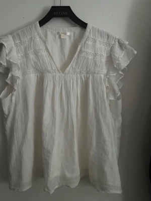 Vit blus med volangärmar - Söt vit blus med v-ringning och volangdetaljer vid ärmarna. Blusen har ett lätt strukturerat tyg och fina broderade detaljer upptill. Perfekt för en fräsch och luftig look.