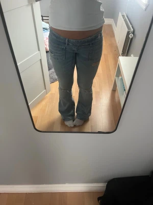 Ljusblå bootcut jeans med slitningar - Säljer ett par ljusblå bootcut jeans med slitningar på låren och knäna. Jeansen har låg midja och klassiska fickor fram och bak. Perfekta för en avslappnad och trendig look. Materialet är denim med lite stretch för extra komfort.
