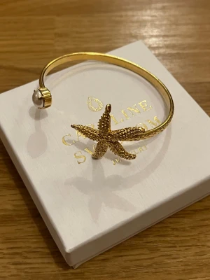 Guldarmband med sjöstjärna och pärla - Snyggt guldarmband från Caroline Svedbom med en detaljrik sjöstjärna i mitten och en vit pärla på ena änden. Armbandet är öppet och har en elegant, havsinspirerad design som verkligen sticker ut.