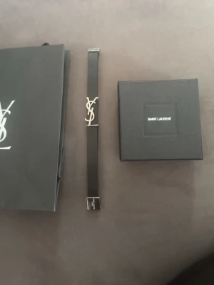 Saint Laurent svart armband med logga - Snyggt svart armband från Saint Laurent med den ikoniska YSL-loggan i silverfärgad metall. Armbandet har en stilren och minimalistisk design med justerbart spänne. Perfekt accessoar för dig som gillar exklusiva detaljer och mode.