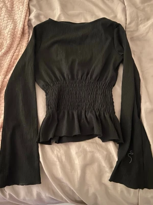 Söt tröja från Gina💕 - Snygg svart topp med lång ärm och smockad midja som ger en figurnära passform. Toppen har volangdetalj nedtill och är tillverkad i ett lätt, strukturerat tyg. Perfekt att styla med jeans eller kjol för en trendig look.
