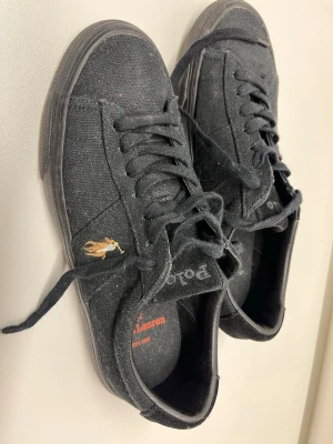 Svarta sneakers från Polo Ralph Lauren - Stilrena svarta sneakers från Polo Ralph Lauren med klassisk låg profil och canvasmaterial. Skorna har svarta snören, beige sula och den ikoniska broderade polospelaren i gult och vitt på sidan. Perfekta för en avslappnad och trendig look. Aldrig använda pga fel storlek 
