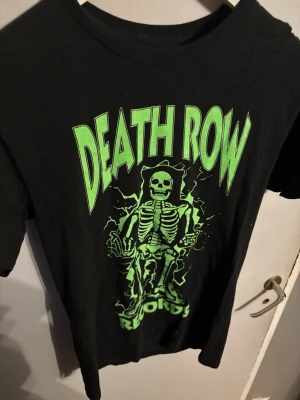 Death Row Records svart t-shirt - Svart t-shirt från Death Row Records med stort grönt tryck av ett skelett och texten 'Death Row Records' på framsidan. Klassisk rund hals och korta ärmar. Perfekt för dig som gillar streetwear och vill sticka ut med en ikonisk design.