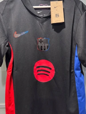 Svart FC Barcelona matchtröja Nike - Snygg svart FC Barcelona matchtröja från Nike med klubbmärke på bröstet, röd och blå detalj på sidorna samt Spotify-logga framtill. Tröjan har korta ärmar och V-ringning, tillverkad i lätt och ventilerande material som passar perfekt till fotboll eller träning.
