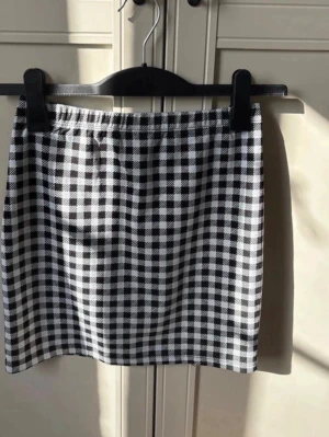 Svartvit rutig minikjol med slits - Säljer en svartvit rutig kortkjol med liten slits på sidan. Kjolen har en rak passform och elastisk midja för skön passning. Perfekt att styla med en enkel topp eller stickad tröja. Materialet känns mjukt och följsamt.