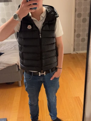 Svart dunväst från Moncler med huva - Snygg svart dunväst från Moncler med huva och dragkedja framtill. Västen har Moncler-logga på bröstet och quiltad design som ger en modern look. Perfekt att slänga över en hoodie eller t-shirt för extra stilpoäng.