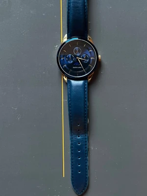 Blå klocka från Filippo Loreti - Stilren klocka från Filippo Loreti med marinblått läderarmband och guldfärgat boett. Urtavlan är blå med flera små ur och gulddetaljer. Baksidan har en graverad illustration av en byggnad. Perfekt accessoar för dig som gillar klassisk men modern stil.