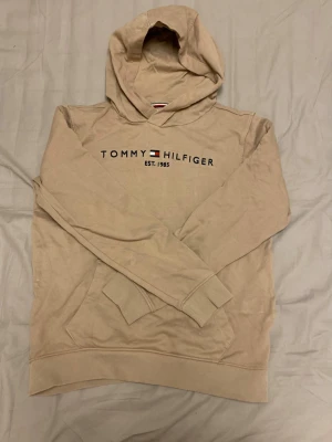 Beige hoodie från Tommy Hilfiger - Snygg beige hoodie från Tommy Hilfiger med klassisk logga och text på bröstet. Hoodien har huva, magficka och ribbade muddar vid ärmslut och nederkant. Tillverkad i mjukt bomullsmaterial som känns skönt mot huden. Perfekt för en avslappnad och trendig look.