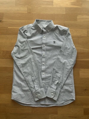 Randig skjorta från Giordano slim fit - Snygg vit och grå randig skjorta från Giordano i slim fit-modell. Skjortan har klassisk krage, långa ärmar och knappar framtill. Tillverkad i bomull och har en liten broderad logga på bröstet. Perfekt för en clean och stilren look.