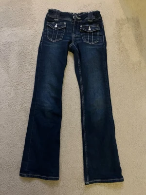 Mörkblå bootcut jeans med fickdetaljer - Snygga mörkblå bootcut jeans med kontrastsömmar och dekorativa fickor framtill med lock och knappar. Jeansen har klassisk femficksdesign och är tillverkade i denim med lätt stretch. Perfekta för en avslappnad och trendig look.