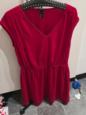 Röd kortklänning från H&M - Snygg röd kortklänning från H&M med v-ringning och resår i midjan. Klänningen är ärmlös och har en enkel, avslappnad passform som passar perfekt till sommaren. Tillverkad i ett mjukt och lätt material.