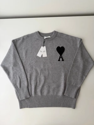 Grå stickad tröja från Ami Paris - Snygg grå stickad tröja från Ami Paris med svart hjärta och bokstaven A broderat på bröstet. Tröjan har rund halsringning, ribbade muddar och är tillverkad i mjukt material som känns lyxigt. Perfekt för dig som gillar stilrena plagg med en twist.