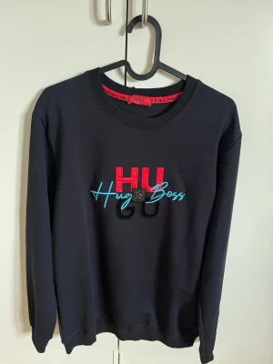Svart sweatshirt från Hugo Boss - Snygg svart sweatshirt från Hugo Boss med broderad logga i rött, blått och svart på bröstet. Rund halsringning och långa ärmar. Perfekt för en casual och trendig look. Mjuk bomullskvalitet och bekväm passform.