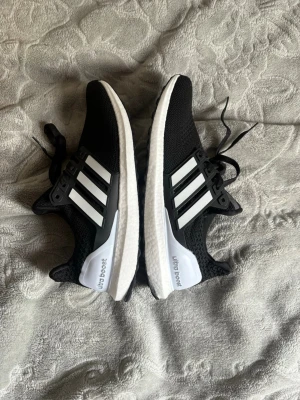Adidas Ultraboost svarta sneakers - Svarta Adidas Ultraboost sneakers med klassiska vita ränder på sidorna och vit sula. Ovandelen är i mesh för bra andningsförmåga och komfort. Snygg och sportig design med snörning och diskret logga på hälen. Perfekta för dig som gillar streetwear och sportig stil. Helt nya oanvända storlek 44