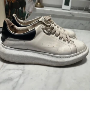 Vita Alexander McQueen sneakers - Stilrena vita sneakers från Alexander McQueen med svart detalj på hälen och tjock sula. Klassisk låg modell med vita skosnören och diskreta hål på sidan för ventilation. Perfekta för dig som gillar en clean och trendig look. Storlek 45