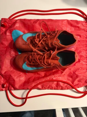 Nike Mercurial röda fotbollsskor elite - Säljer ett par Nike Mercurial fotbollsskor i rött med turkosa detaljer och snörning. Skorna har en låg profil och är tillverkade i ett lätt syntetmaterial för bra bollkontroll. Perfekta för dig som vill ha snabbhet och stil på planen.