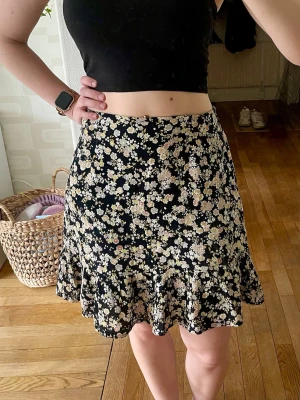 Svart blommig minikjol - Supersöt svart kortkjol med blommigt mönster i svart och gult. Kjolen har en lätt utställd form och volang nedtill som ger en flowy känsla. Perfekt för vår och sommar, matcha med en enkel topp för en fräsch look.
