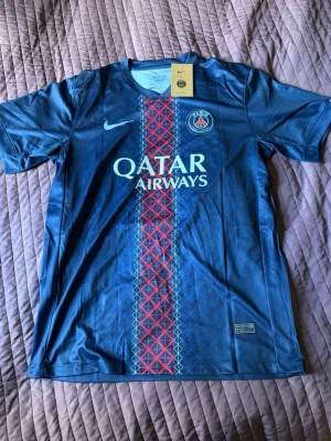 PSG matchtröja Dembélé #10 Nike blå - Snygg blå PSG matchtröja från Nike med Qatar Airways-tryck på bröstet och O. Dembélé #10 på ryggen. Tröjan har korta ärmar, V-ringning och ett rött och vitt mönster längs mitten. Perfekt för fotbollsfans som vill sticka ut på läktaren eller planen.
