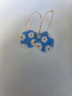 Blå handgjorda örhängen med prästkragemotiv - Somriga örhängen med prästkragemotiv på blå botten. Sitter på guldiga hoops av rostfritt stål med en diameter på 2,5 cm. Gjorda för hand av mig i polymerlera. 