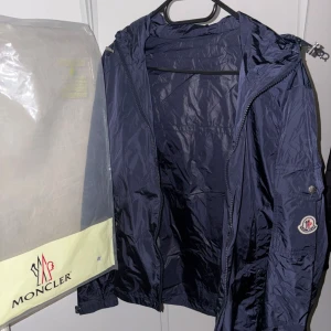 Mörkblå vindjacka från Moncler - Snygg mörkblå vindjacka från Moncler med huva och dragkedja framtill. Jackan har Moncler-logga på ärmen, praktiska fickor med knappar och justerbar snörning vid huvan. Materialet är lätt och glansigt polyester, perfekt för blåsiga dagar. Passar både M och S