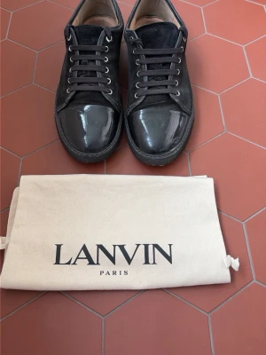 Svarta Lanvin sneakers i mocka och lack - Snygga svarta sneakers från Lanvin med ovandel i mocka och lackade tåpartier. Klassisk låg modell med svarta skosnören och metallöljetter. Kommer med original dustbag från Lanvin Paris. Perfekta för dig som gillar stilrena och exklusiva sneakers.