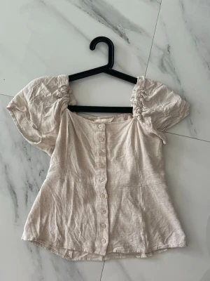 Beige blus med puffärmar och knappar - Säljer en söt beige blus med puffiga korta ärmar och knappar hela vägen framtill. Blusen har en lätt strukturerad yta och en feminin form som framhäver midjan. Perfekt för dig som gillar romantiska detaljer och enkel stil.