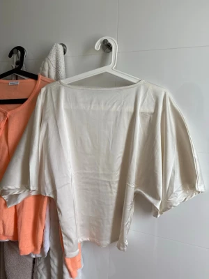 Vit topp med vida ärmar - Säljer en vit topp med supervida, nästan fladdriga ärmar och rund halsringning. Materialet känns mjukt och har ett lätt glansigt utseende, vilket ger toppen en lyxig känsla. Perfekt för dig som gillar avslappnad och trendig stil.
