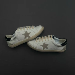 Vita Golden Goose med svart detalj - Stilrena vita sneakers från Golden Goose med svart hälparti, beige och svart sula. Klassisk låg modell med vita skosnören och diskreta håldetaljer på sidan. Perfekta för en clean och avslappnad look.