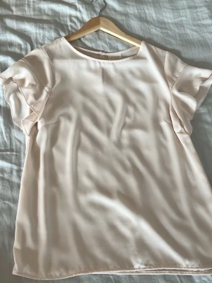 Ljusrosa blus med volangärmar - Söt ljusrosa blus i mjukt, glansigt material med korta volangärmar och rund halsringning. Perfekt att styla till jeans eller kjol för en trendig look. Enkel och stilren design utan synliga detaljer eller mönster.
