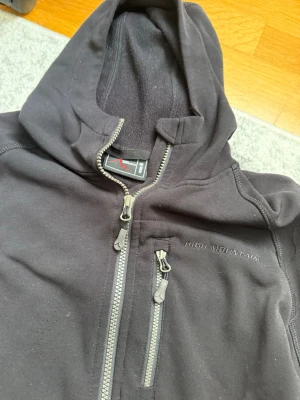 Svart hoodie från High Mountain - Säljer en svart hoodie från High Mountain med dragkedja framtill och tre fickor med dragkedja. Hoodien har broderad logga på bröstet och är gjord i mjukt, fleeceliknande material. Perfekt för chill dagar eller när du vill ha något bekvämt och stilrent. Helt felfri och snygg storlek M den är perfekt  helt enkelt