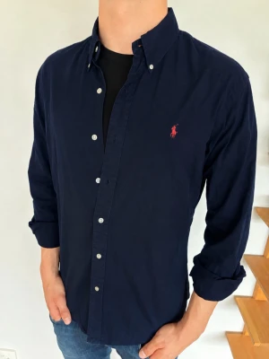 Mörkblå skjorta från Polo Ralph Lauren - Klassisk mörkblå skjorta från Polo Ralph Lauren med knappkrage och den ikoniska röda logobrodyr på bröstet. Skjortan har långa ärmar, vita knappar och är tillverkad i mjuk bomull. Perfekt till jeans eller chinos för en clean look.