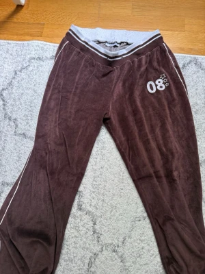 Bruna mjukisbyxor med tryck - Mysiga bruna mjukisbyxor i velour med bred, ribbad midjeresår i grått och vita detaljer längs sidorna. På vänster lår finns ett vitt tryck med siffran 08 och små stjärnor. Perfekta för chill dagar hemma eller till träningen. Passar även M