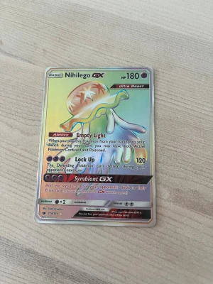 Nihilego GX Pokémonkort Ultra Beast - Säljer ett holografiskt Nihilego GX Ultra Beast Pokémonkort med 180 HP. Kortet har färger i regnbågsskimrande toner och detaljer i silver. Det är på engelska och har attacker som Lock Up och Symbiont GX. Perfekt för samlare eller Pokémonfans.