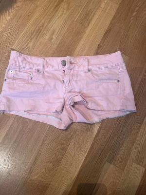 Ljusrosa jeansshorts med rå kant - Säljer ett par ljusrosa jeansshorts med råa kanter och klassisk femficksdesign. Shortsen har knapp och dragkedja framtill samt snygga bakfickor med broderad detalj. Perfekta för sommaren och riktigt sköna att bära.