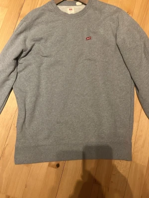 Grå sweatshirt från Levi's - Klassisk grå sweatshirt från Levi's med rund halsringning och liten röd Levi's-logga på bröstet. Tröjan har en enkel och stilren design med ribbade muddar vid ärmslut och nederkant. Perfekt för en avslappnad och snygg look.