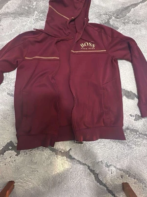 Vinröd hoodie - Säljer en vinröd hoodie med dragkedja och huva. Hoodien har guldfärgade detaljer och logga på bröstet samt ribbade muddar vid ärmslut och nederkant. Perfekt för en chill och stilren look.