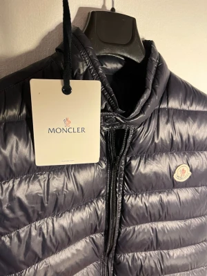 Svart dunväst från Moncler - Snygg svart dunväst från Moncler med quiltad design och hög krage. Västen har dragkedja framtill, två fickor med dragkedja och Moncler-logga på bröstet. Tillverkad i glansigt material som ger en modern look. Storlek 3 vilket motsvarar S/M. Dragkedjan har jag bytt ut eftersom de lätt går sönder på denna modell.
